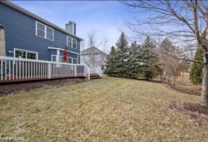 Backyard view of a blue house with a deck, surrounded by trees and grass, showcasing outdoor space relevant to real estate services in Harthside, Illinois.